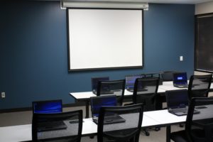 an image of a classroom with tables, chairs, a screen, and laptops on the tables